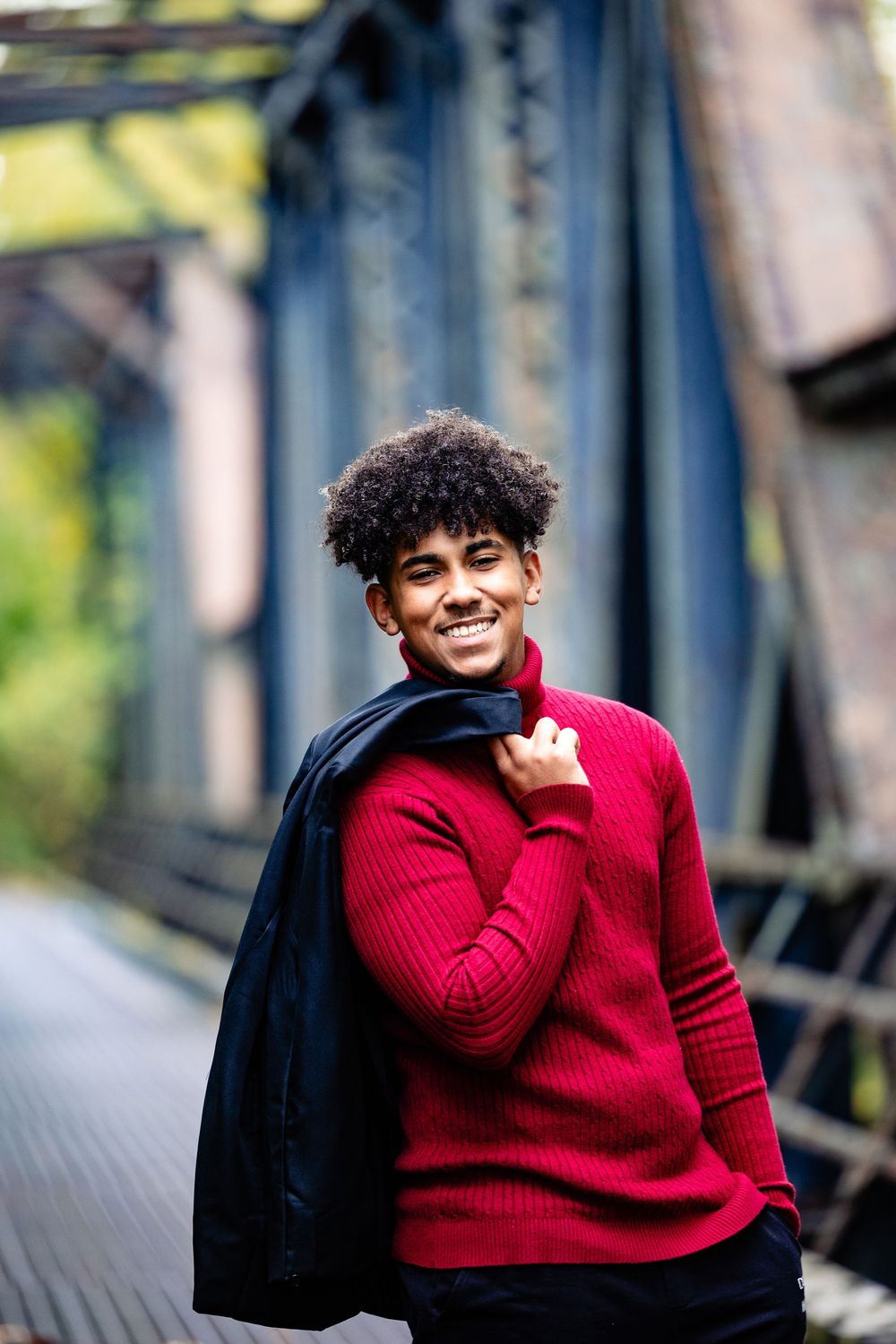 Young Black man with curly hair smiles confidently, wearing a red sweater and holding a jacket over his shoulder on a bridge.