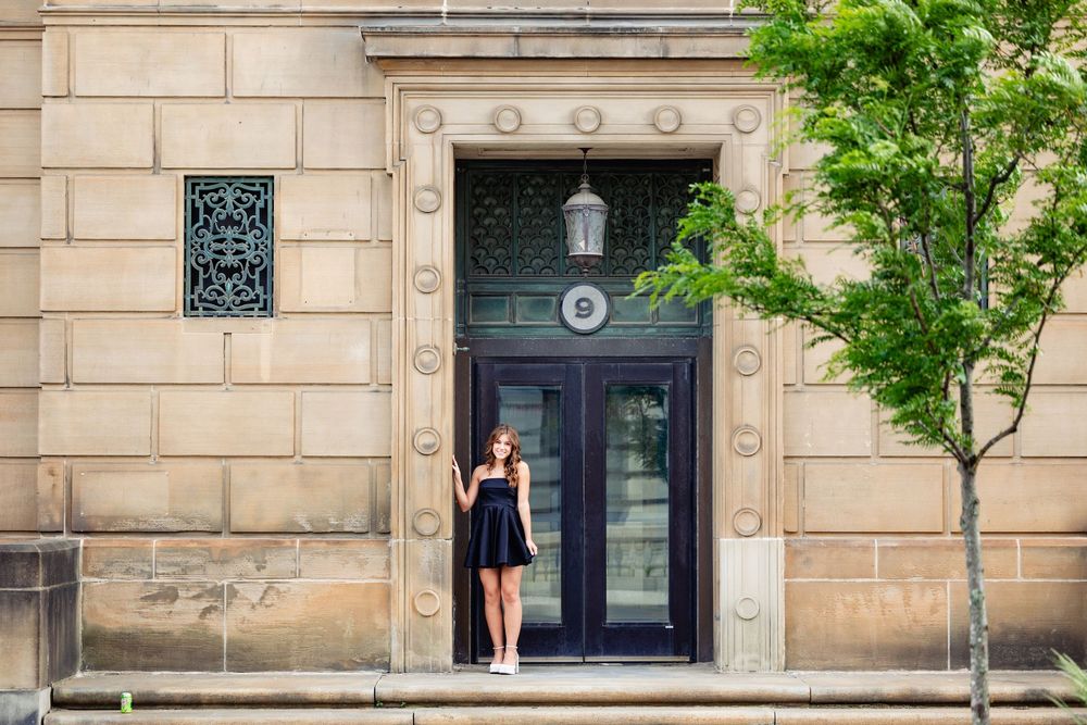Woman in black dress standing by ornate stone building entrance with decorative door and green trees.