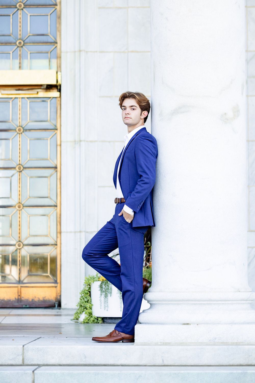 Young man in blue suit leaning against white marble column near ornate gold door.