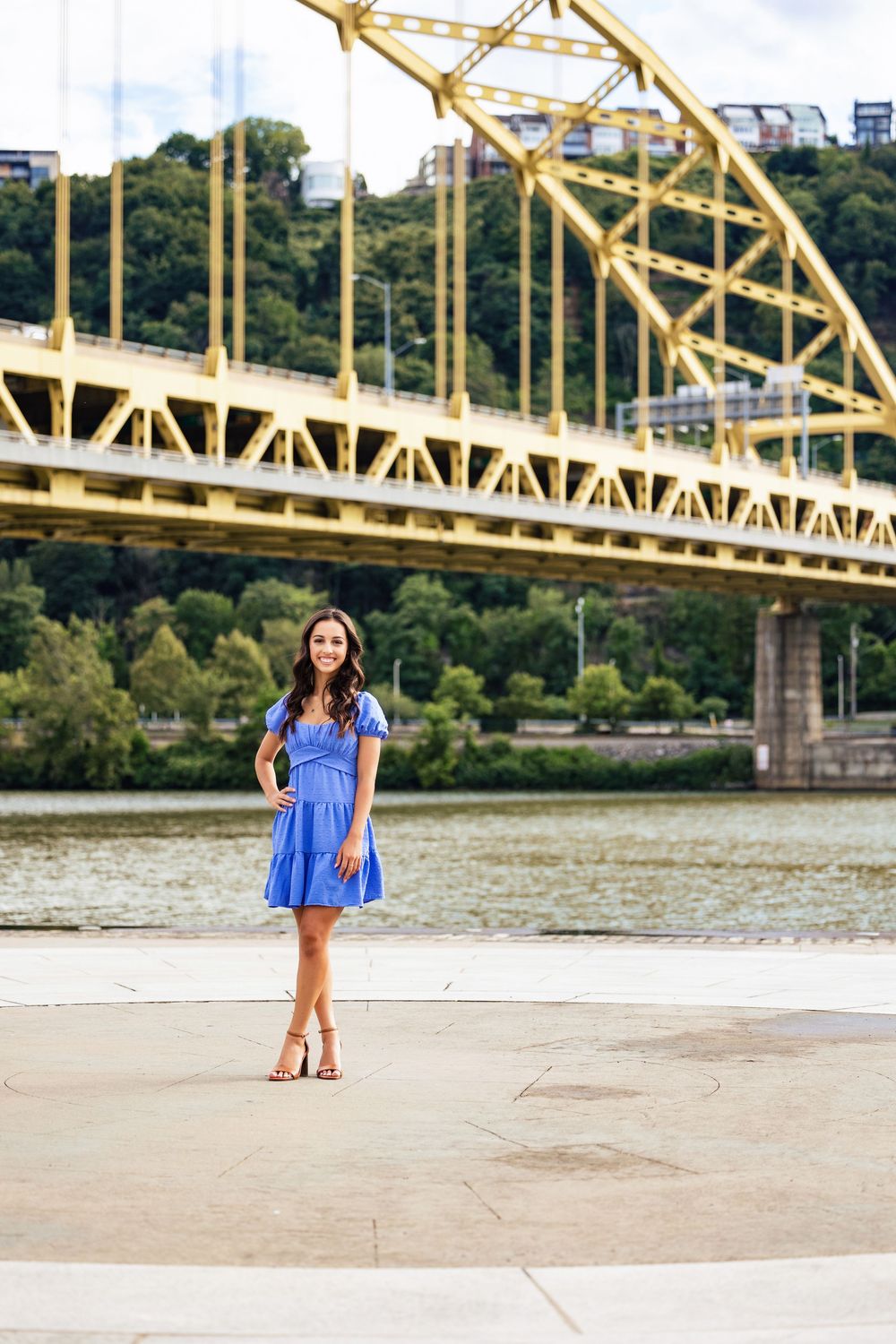 Young woman in blue dress posing by Pittsburgh river with yellow Fort Pitt Bridge in background.