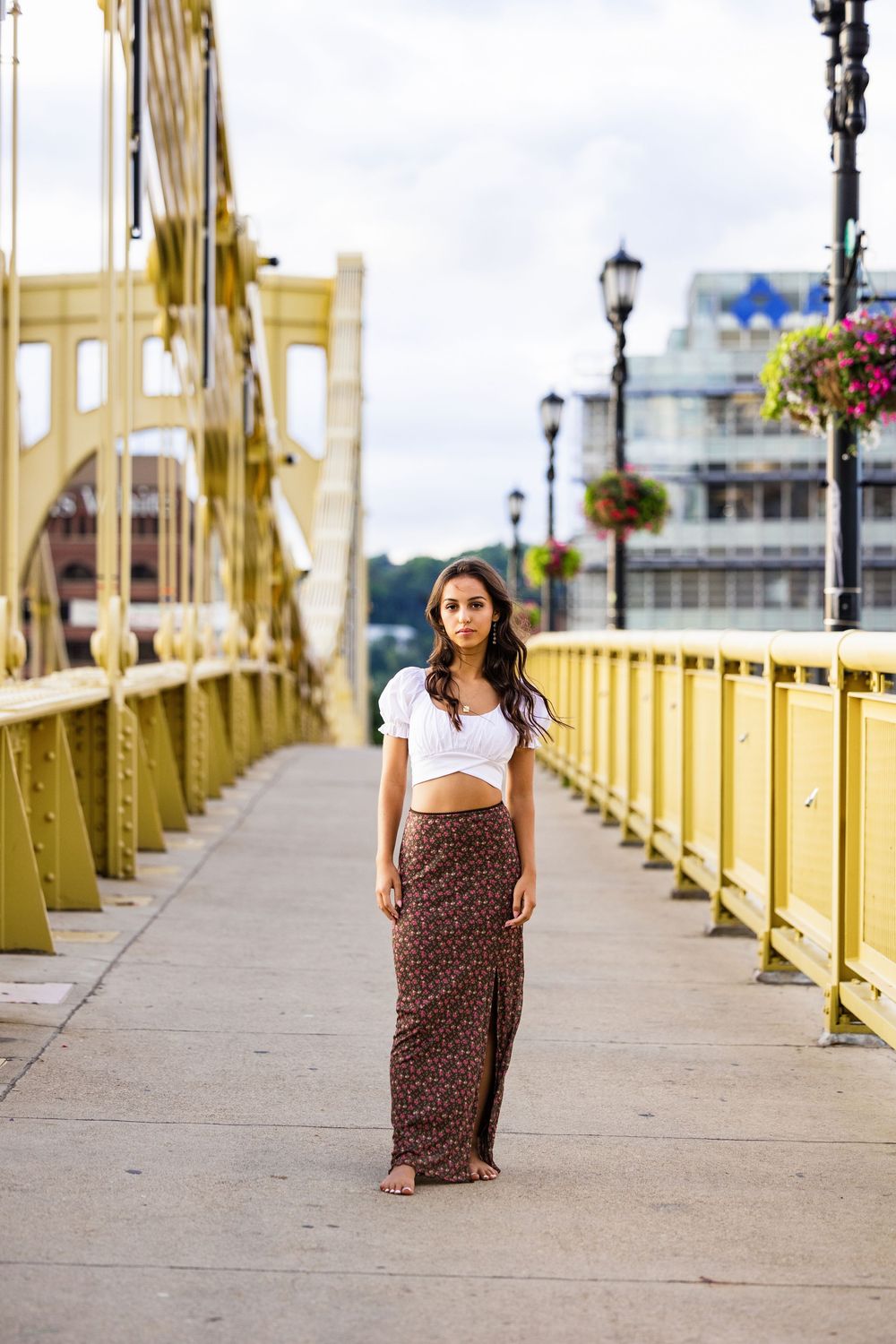 Young woman in white crop top and floral maxi skirt standing on a yellow bridge with city views.