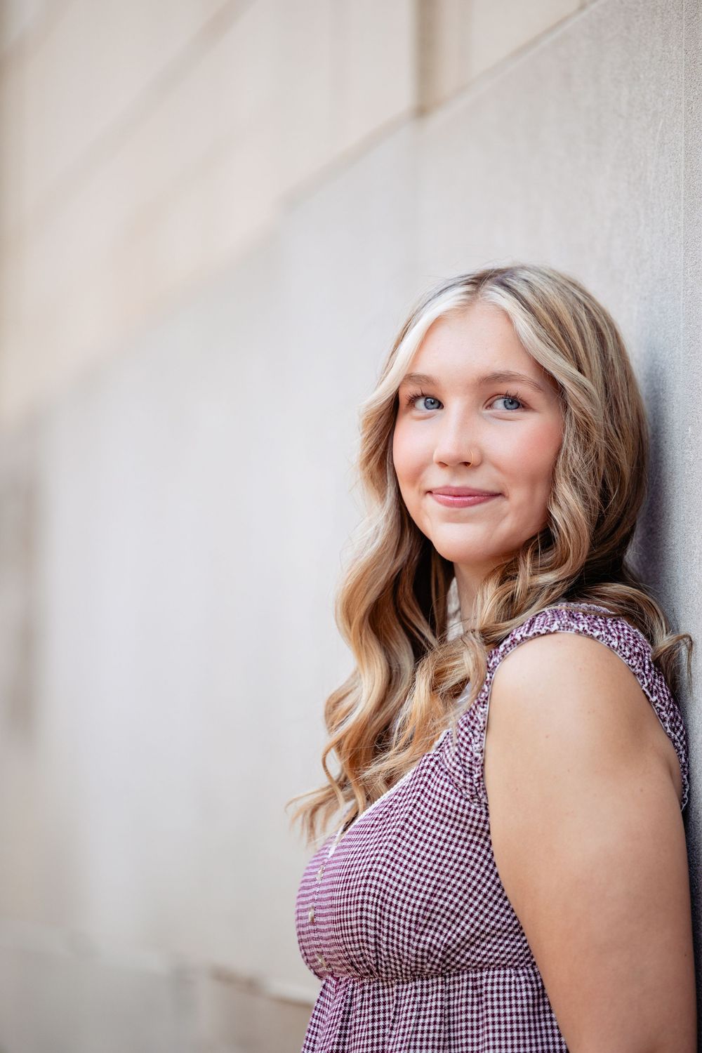 Young blonde woman in a purple dress leaning against a wall, smiling softly at the camera.