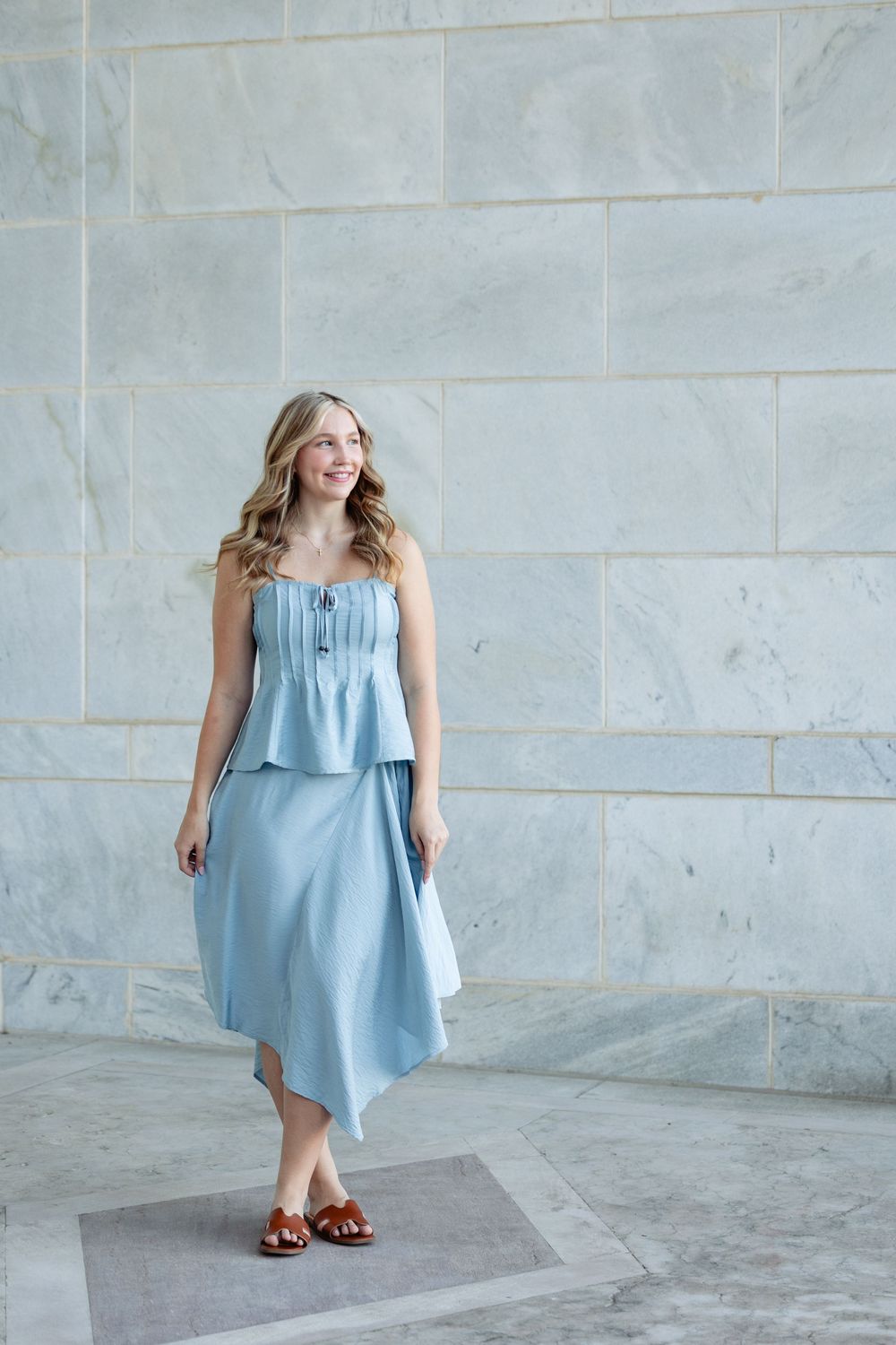 Woman in light blue strapless top and midi skirt with brown sandals posing against marble wall.