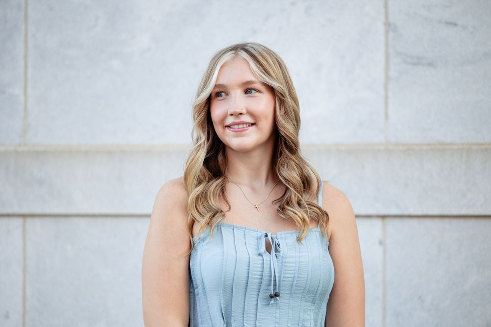 Smiling young blonde woman in a light blue sleeveless top posing outdoors against a marble wall.