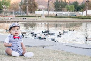 Baby in formal attire sits near a pond with ducks swimming in the background.