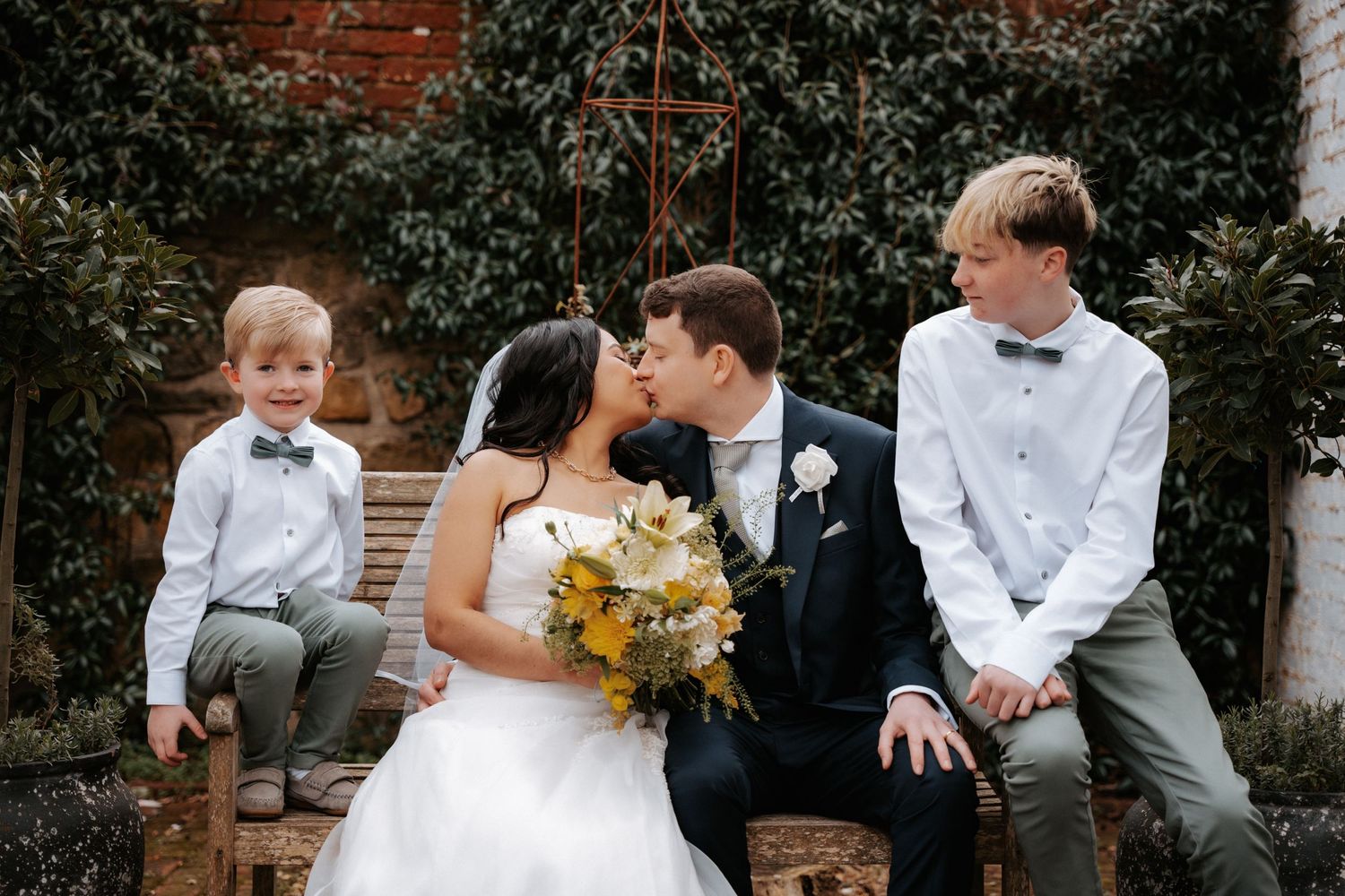 Wedding ceremony guests sit on wood benches in an outdoor garden setting decorated with white flowers and greenery.
