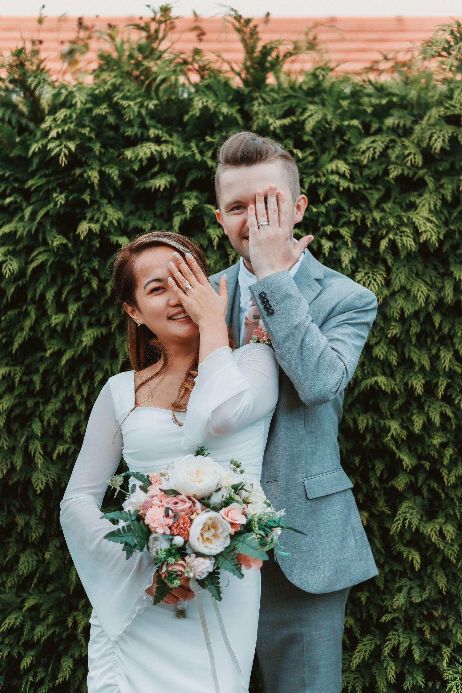 Wedding couple shares candid laughing moment in front of garden hedge.
