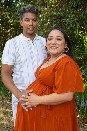Couple embracing outdoors with person wearing orange dress against green foliage background.