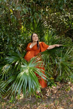 Person in orange dress poses playfully among tropical palm fronds in garden setting.