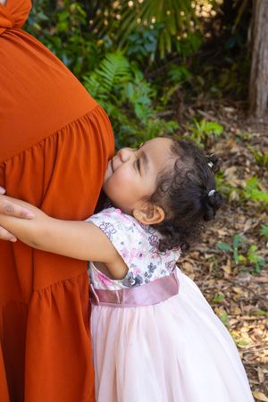 Young girl in a pink dress hugs someone wearing a rust-colored dress in an outdoor setting.