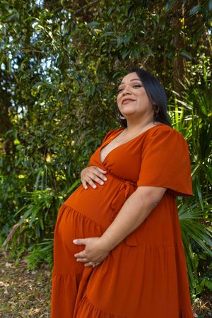 Woman in a rust-colored dress poses outdoors with greenery in the background.