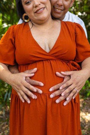 Intimate couple's portrait showing hands cradling a pregnant belly in rust-colored dress.