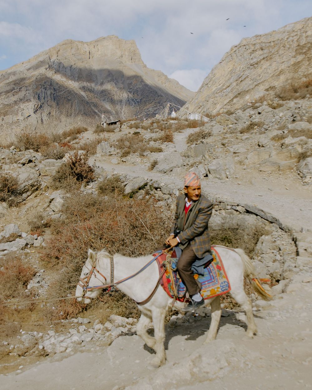 A rider on a white donkey traverses a rocky mountain path with rugged peaks rising in the background.