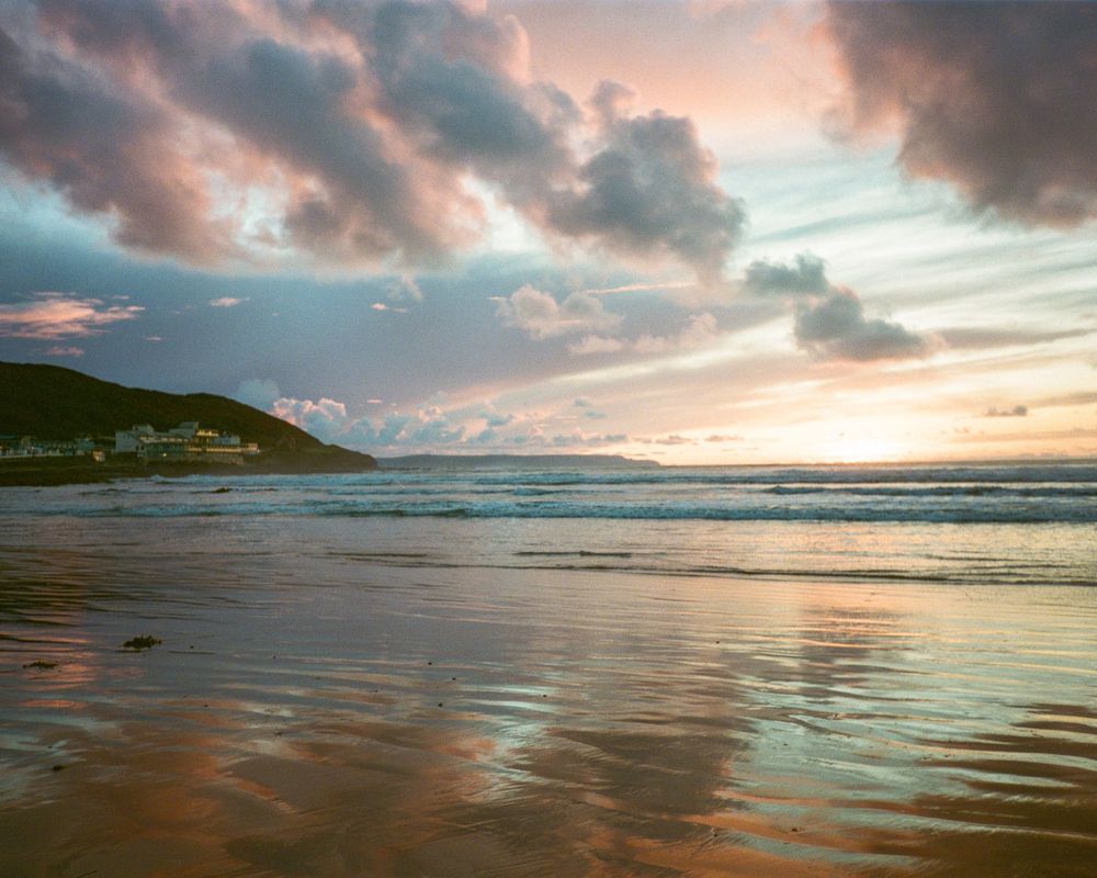 Panoramic view of a dramatic sunset over a beach with pink clouds reflecting on wet sand and gentle waves rolling in.