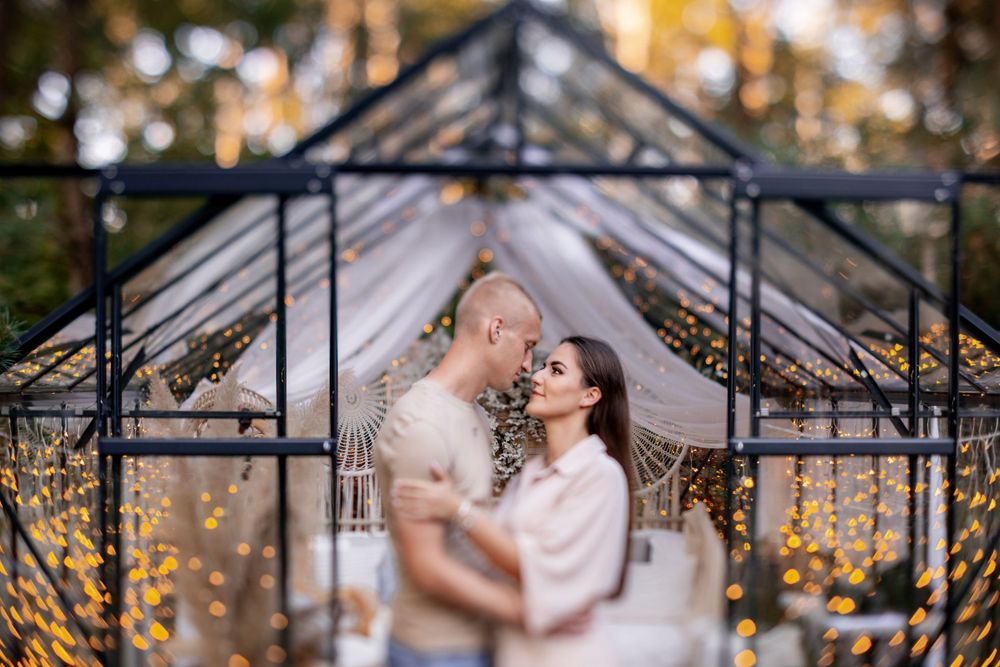 Romantic couple embraces under a greenhouse structure decorated with twinkling fairy lights.