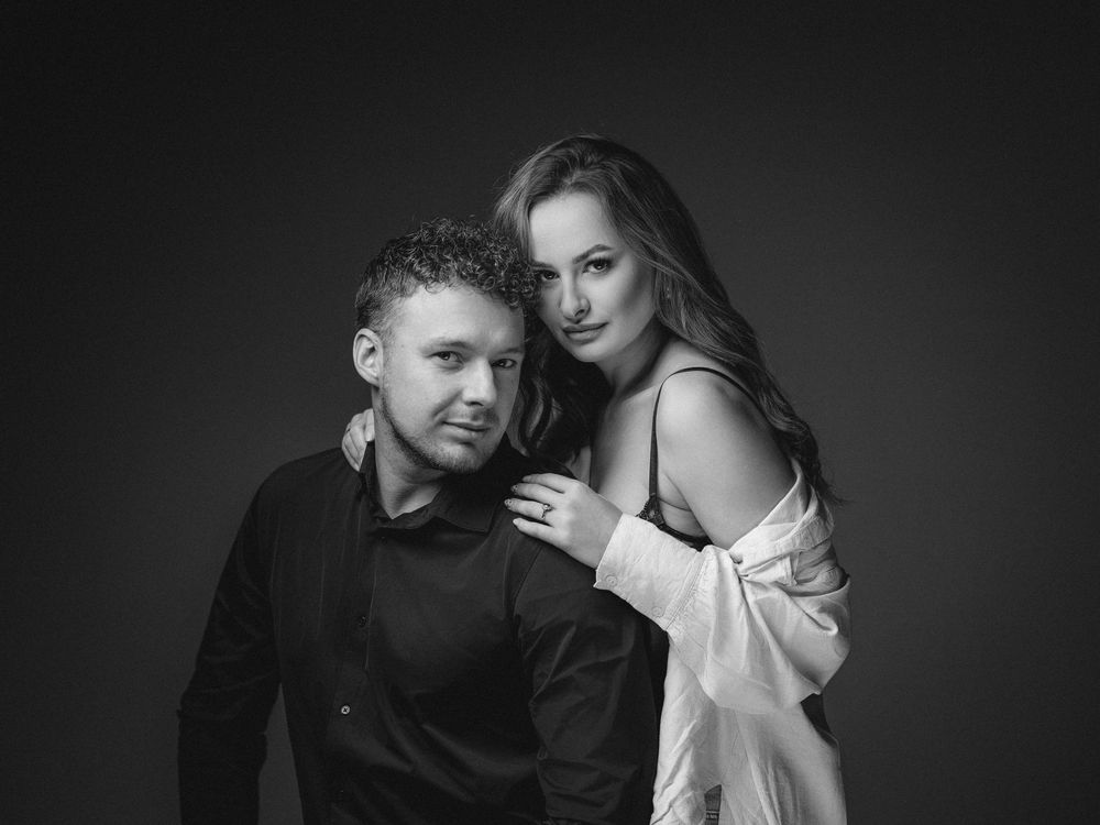 Monochrome studio portrait of a couple in intimate pose against dark background, with dramatic lighting.