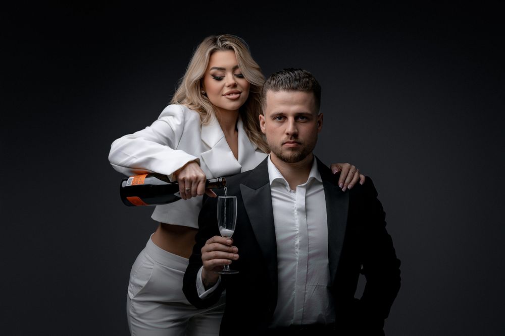 Stylish couple in formal attire posing with champagne against dark background.