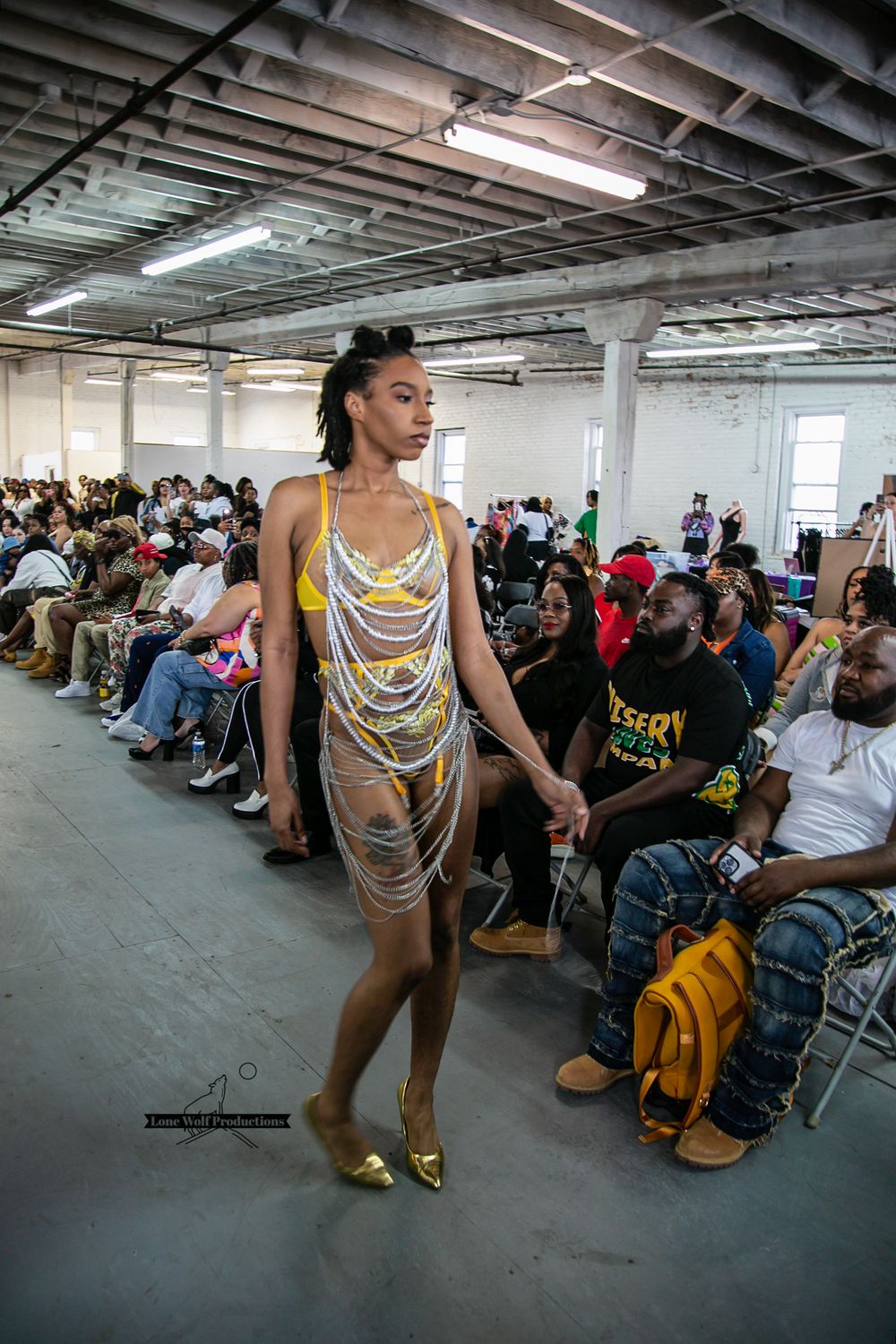 A tall model walks the runway in a striking metallic mesh dress during a warehouse fashion show.
