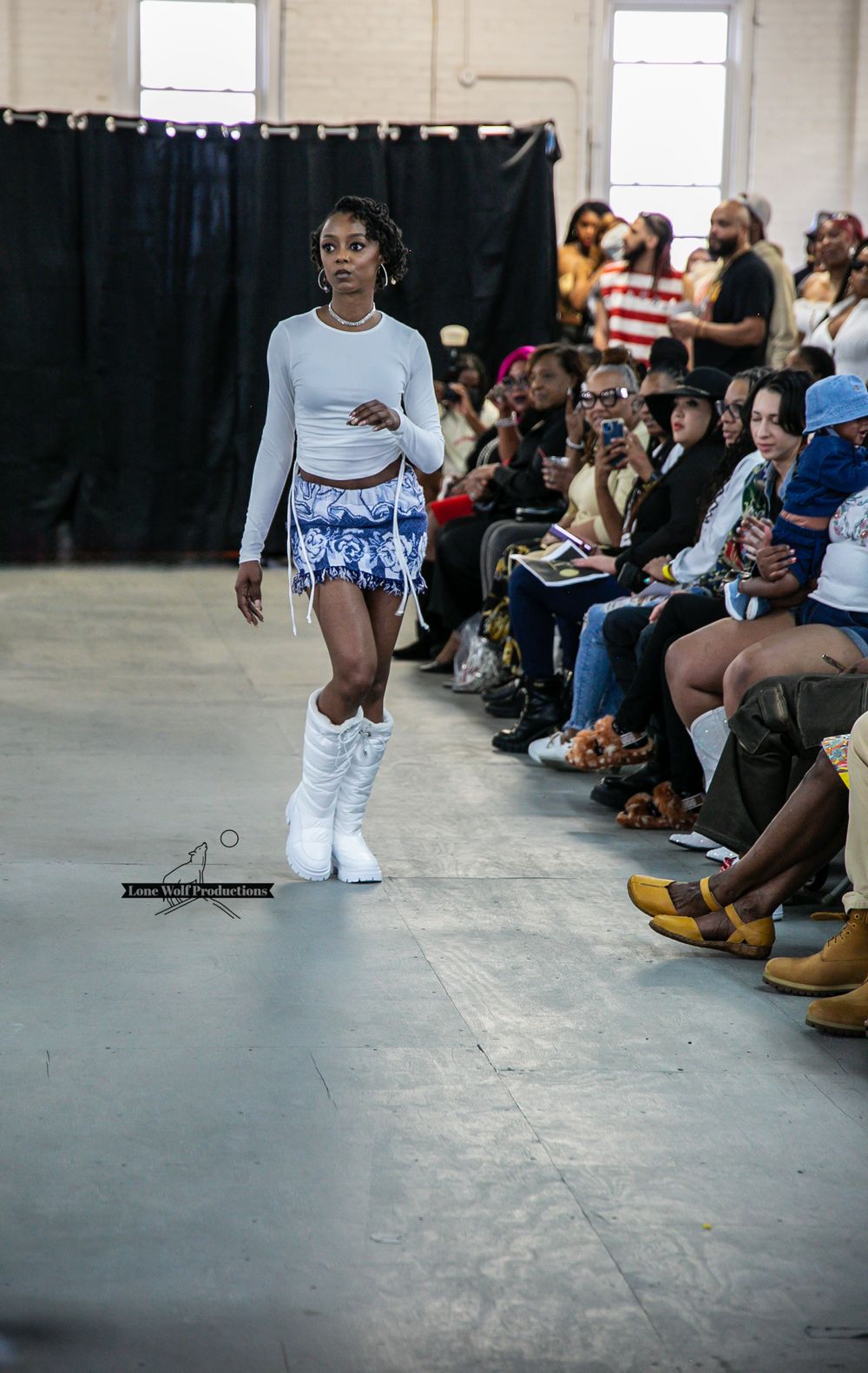 Model walks runway in cropped light blue top, printed shorts and white boots at fashion show.