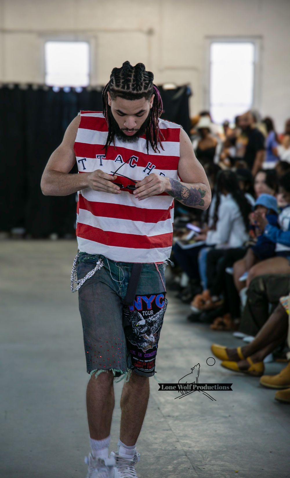 Model walks runway in red and white striped sleeveless shirt and distressed denim shorts during fashion show.