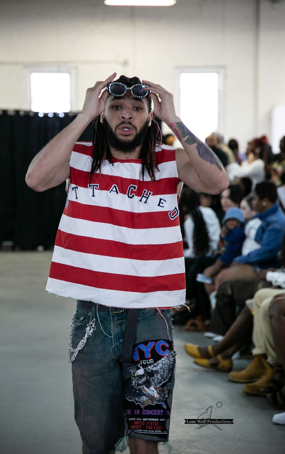 Fashion model adjusts sunglasses while wearing striped tank top and denim shorts on runway.