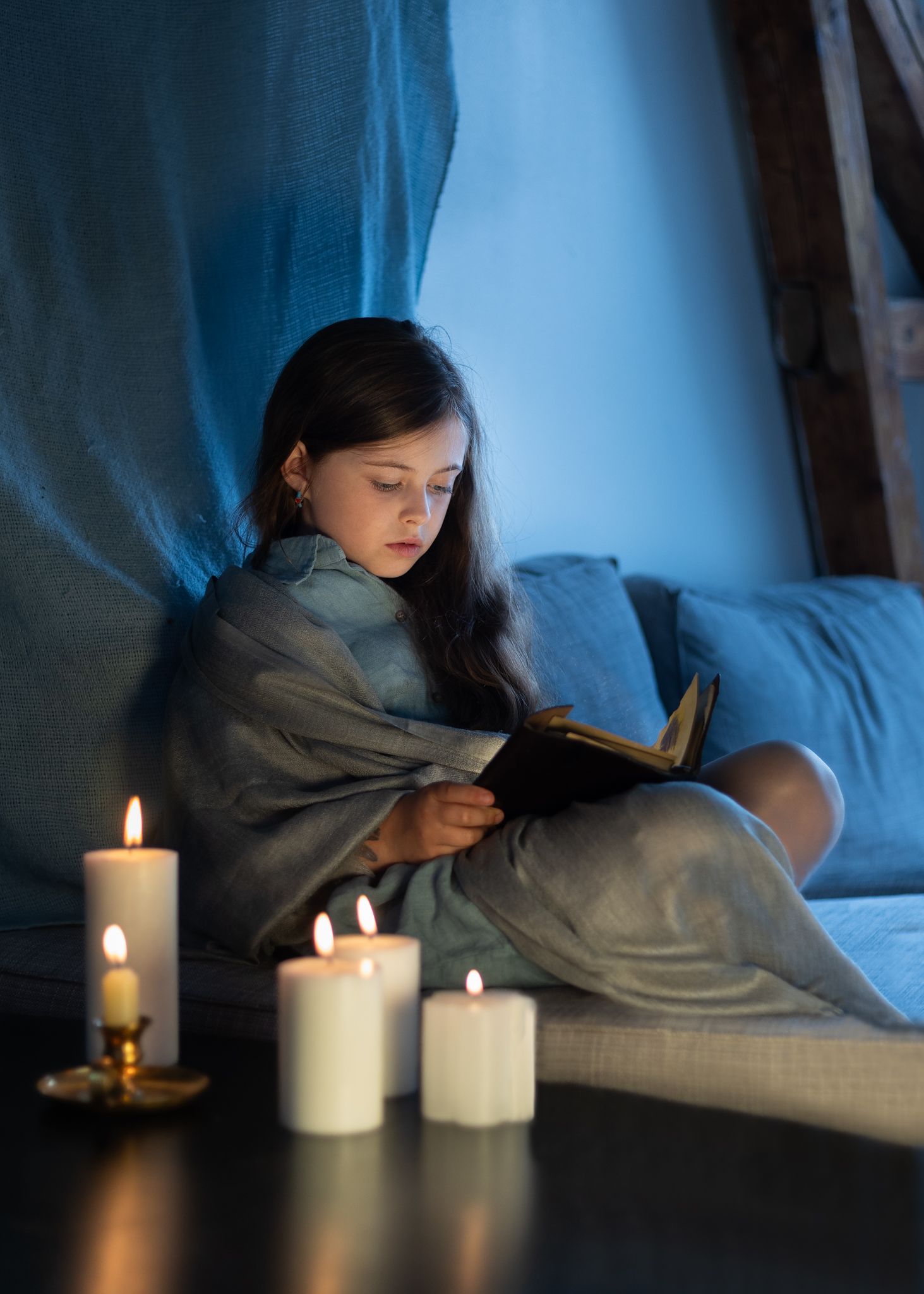 Atmospheric nighttime scene with candles and person reading under blanket in dim blue light.