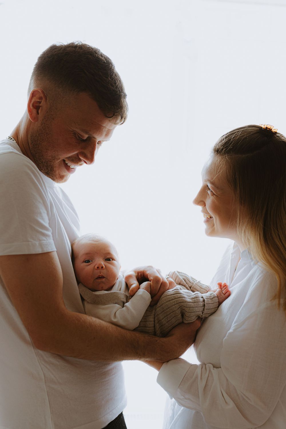 Family in white clothes sharing intimate moment with their newborn by window.