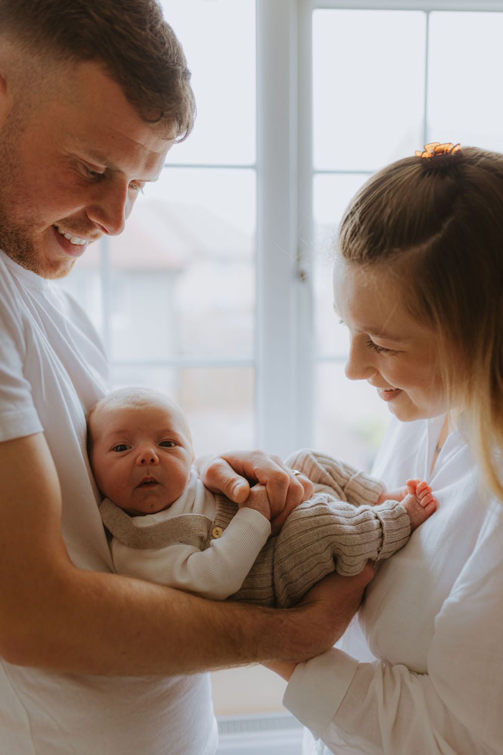 Parents in white shirts holding their newborn baby near bright window.