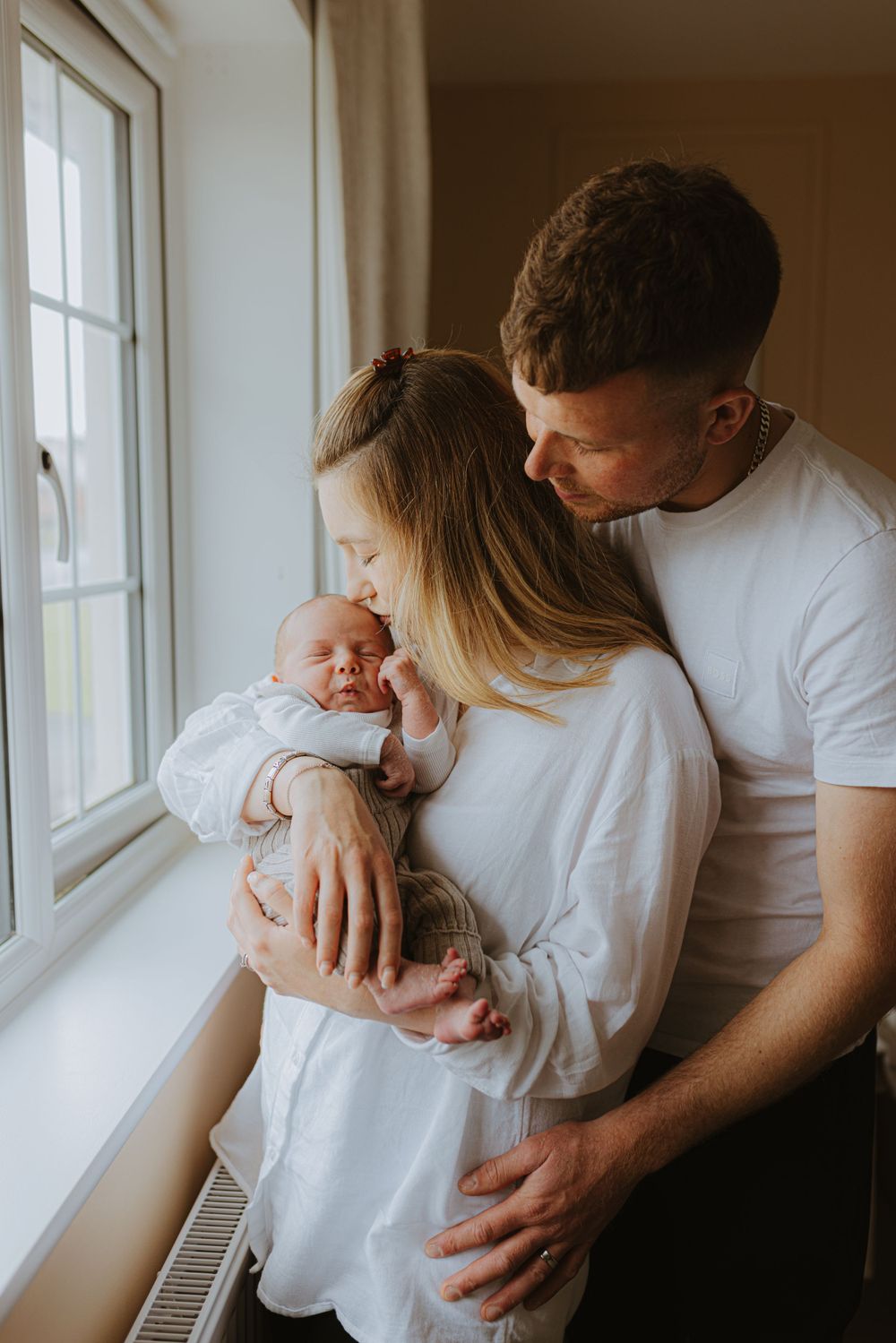 Intimate family moment by window as parents cradle their newborn.