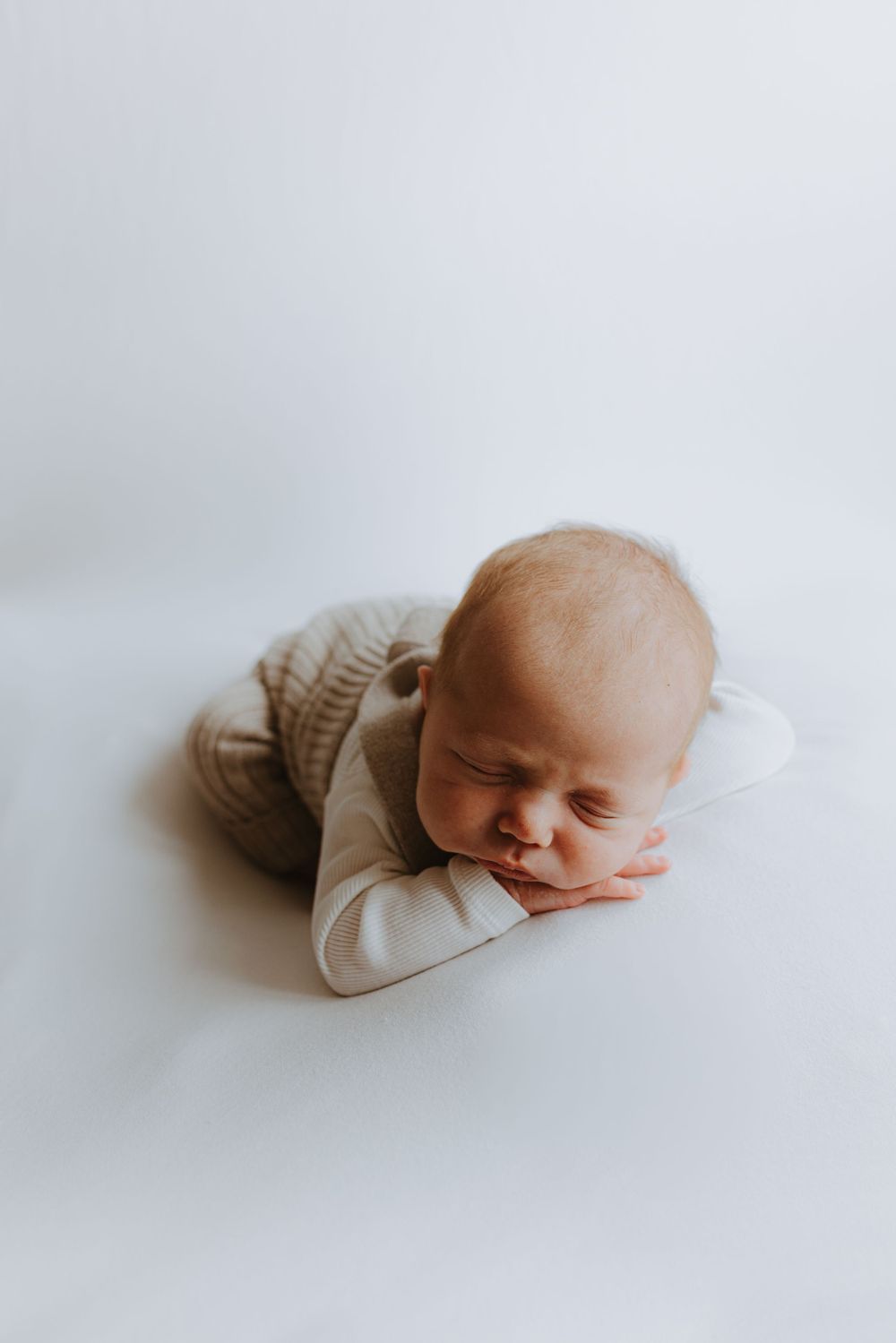 Sleeping newborn in beige outfit lying on white background during photo session.