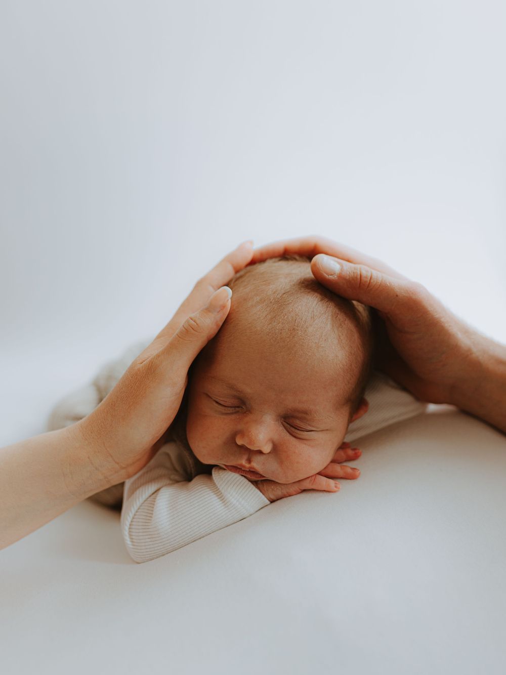 Gentle hands tenderly caress a sleeping newborn baby wrapped in soft white fabric.