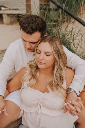 A romantic couple in white outfits share an intimate moment while sitting together on a sandy beach at sunset.