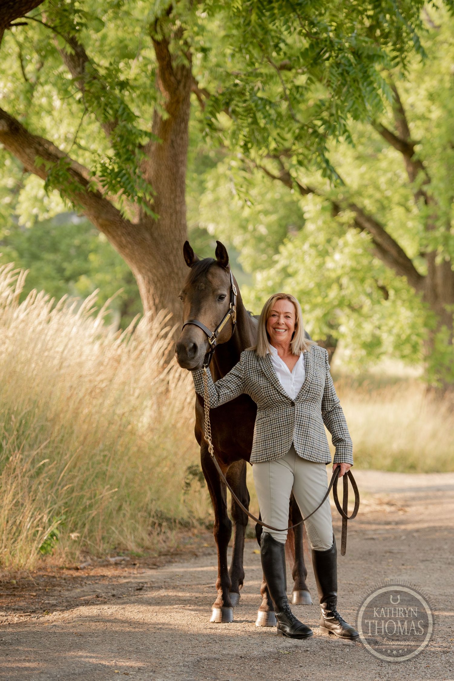 Dark horse and handler wearing a gray jacket pose together on a scenic path under green trees.