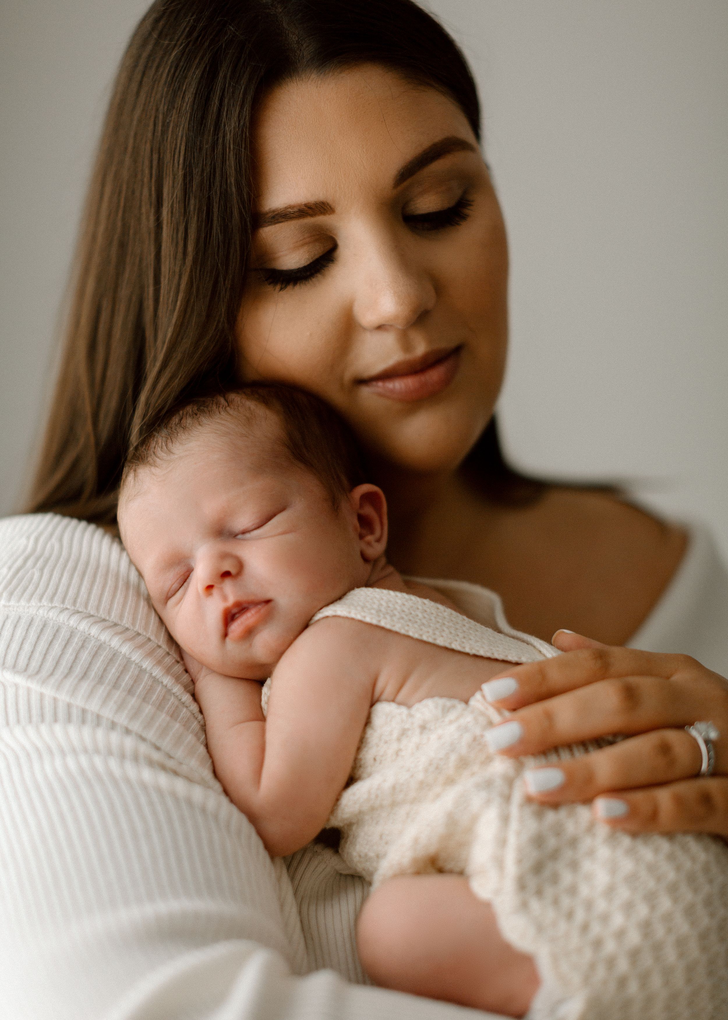 A tender moment between a parent and sleeping newborn baby swaddled in a white knit blanket.