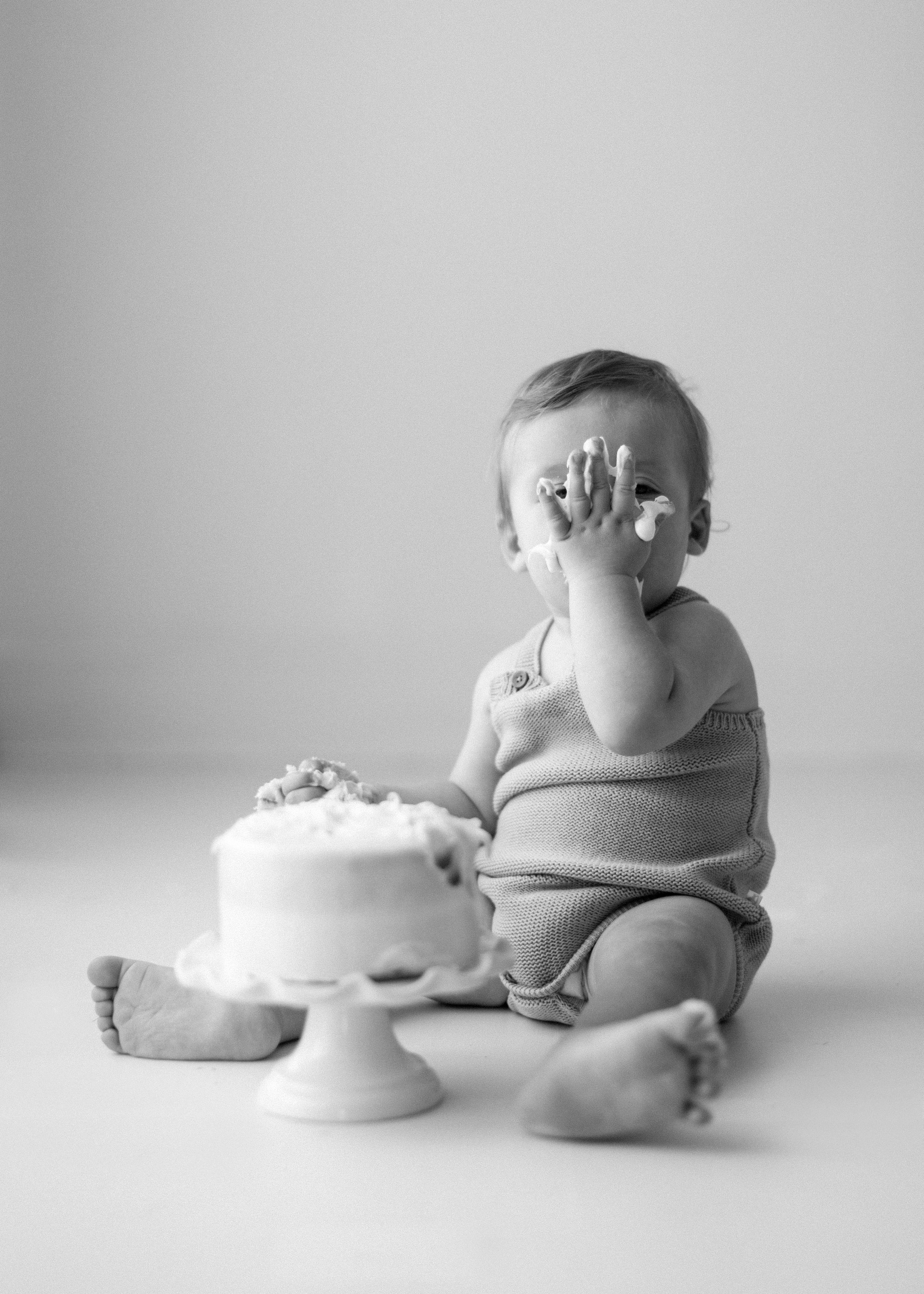 Black and white photo of baby in romper sitting beside white birthday cake on pedestal, covering face with hand.