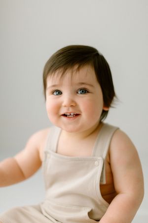 Adorable baby in beige overalls smiling brightly against a light gray background.