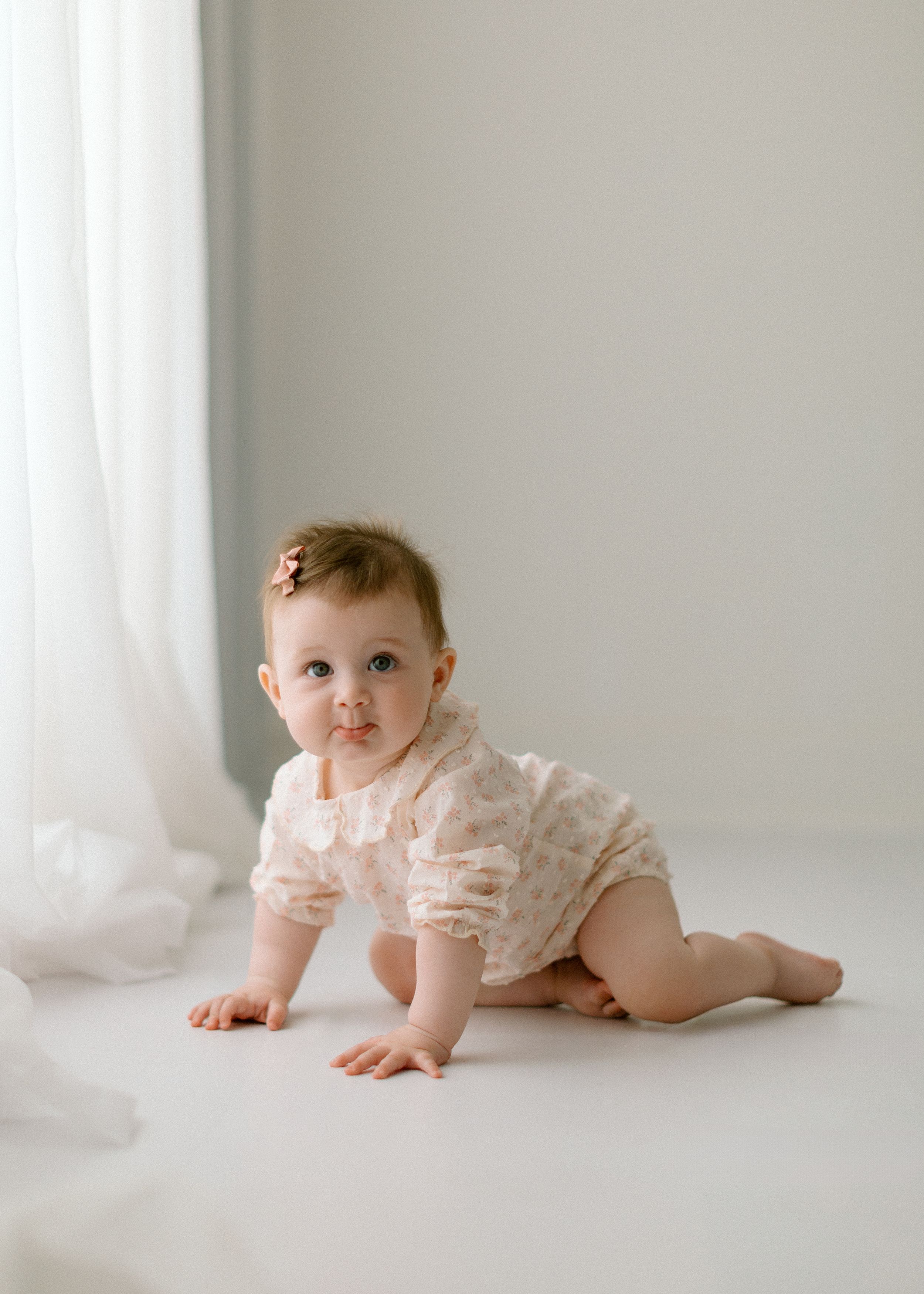 Baby in white lace romper crawls on white floor near sheer curtains in minimalist setting.