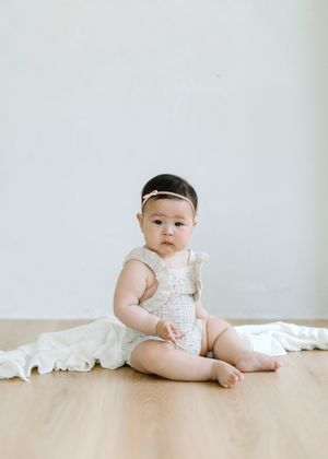 A small child wearing a white lace romper and headband sits on a hardwood floor with white fabric in the background.