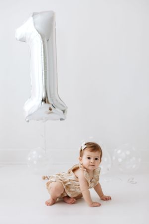 Baby crawls under floating silver number 1 balloon in minimalist setting.