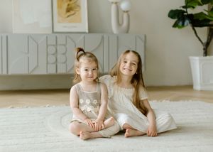 Two young children in light-colored dresses sit together on a white carpet in a bright, airy room.