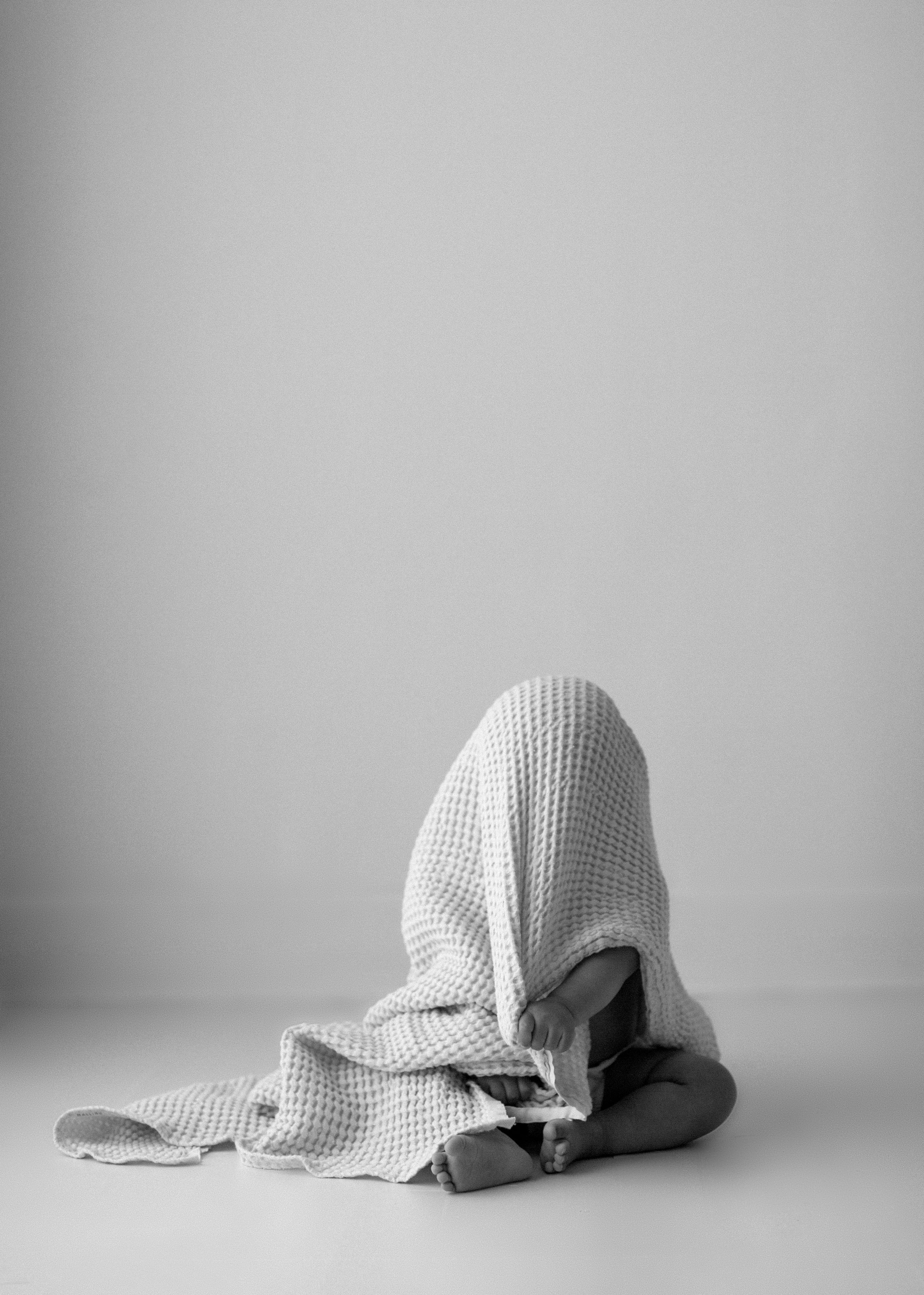 A person covered in a white textured blanket or cloth sits hunched over against a plain gray background.