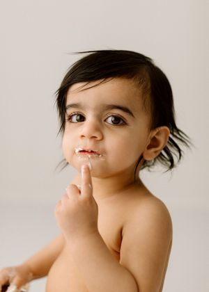 Toddler with dark hair points finger against lips while looking off to the side with playful expression.