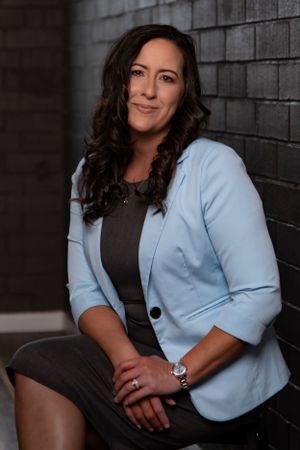 Professional portrait in a light blue blazer and dark outfit against a textured wall.