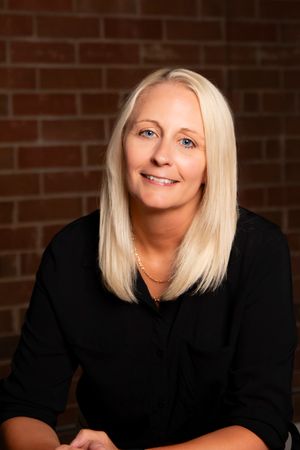 Professional headshot of a woman in black against a brick wall background.
