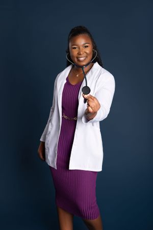Healthcare professional in white coat and purple dress holds stethoscope while smiling against dark blue background personal branding.