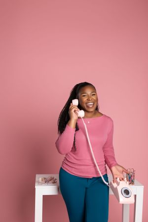 Someone wearing a pink sweater and blue pants talks on a vintage phone while standing against a pink background.
