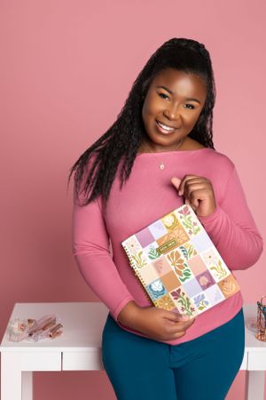 A personal branding photo with a smiling person in a pink sweater holds a colorful floral planner against a pink background.