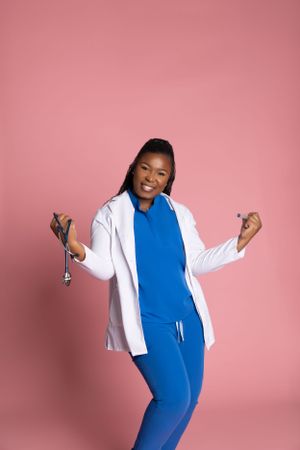 Healthcare professional in blue scrubs and white coat dancing joyfully against pink background with stethoscope.