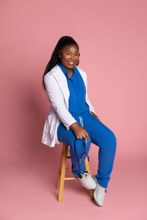 Professional portrait of someone in a blue jumpsuit and white blazer sitting on wooden stool against pink backdrop.