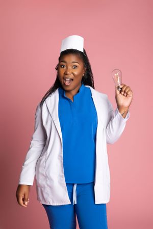 Excited healthcare worker in blue scrubs and white coat holds up a lightbulb against pink background personal branding